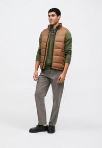 Brown padded vest over a green sweater, gray trousers, and black shoes. The vest has a high collar and front zipper.