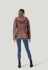 Giacca a vento marrone con cappuccio, caratterizzata da un design trapuntato e una chiusura con zip. Abbinata a jeans blu e stivali neri alla caviglia.