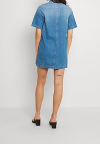 Vestido corto de denim con mangas cortas, degradado de azul claro a oscuro, dos costuras laterales y corte recto. Llevado con sandalias de tacón bloque negro.