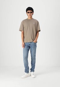 Bruine katoenen t-shirt, lichtblauwe denim jeans, witte sneakers en zwarte zonnebril; staande tegen een effen witte achtergrond.