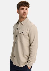Camisa beige de botón, con una tela texturizada, que presenta dos bolsillos en el pecho, botones negros y un cuello tipo americana.