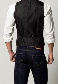 Czarna atłasowa kamizelka o dopasowanym kroju, z regulowanym paskiem z tyłu, noszona na białej koszuli, w połączeniu z ciemnymi dżinsami typu denim z żółtym przeszyciem.