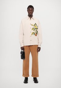 Pārmēra krēmkrāsas krekls ar apelsīnu un zaļu augļu apdruku; pogām aiztaisāms dizains, kombinēts ar brūnām korduroja biksēm un melniem aksesuāriem.