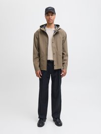 Man i svart keps, beige hoodie, brun jacka, svarta byxor och svarta skor som står mot en enfärgad vit bakgrund.