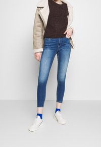 Jeans de mezclilla en azul medio con un corte ajustado, que presentan un ligero desgaste. Combinados con un suéter texturizado de marrón oscuro y zapatillas blancas.