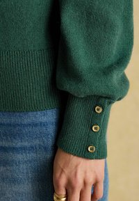 Roheline kudumiga sviiter ribatud varrukatega. Varrukatel on neli kuldset nööpi ja tekstureeritud kangas. Seljas on heledad denimist teksad.