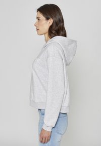 Sweat à capuche gris raccourci avec cordon de serrage, doté de poignets et d'un ourlet côtelés. Porté avec un jean en denim bleu clair, présenté en vue de profil.