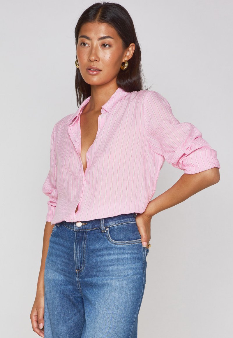 Chemise rose à boutons avec de fines rayures blanches verticales, manches retroussées et col légèrement ouvert, associée à un jean bleu taille haute.
