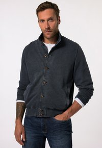 Dark blue button-up jacket with a high collar, wooden buttons, and ribbed cuffs; worn over a white shirt, paired with blue denim jeans.