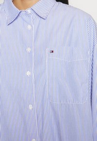 Chemise à rayures verticales bleues et blanches avec une poche avant ornée d'un petit écusson logo rouge, blanc et bleu.