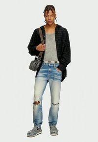 Jauns vīrietis ar pīts matiem, ģērbies melnā rūtainā krekliņā virs pelēka bez Piedurkņu krekla, saplēstos zilos džinsos, sporta apavos un ar pārsega somu.