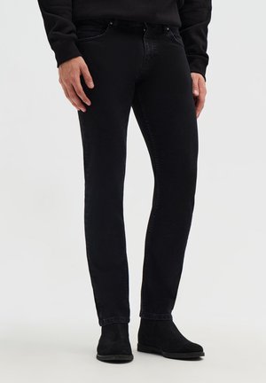 Uomo che indossa jeans neri aderenti con stivali neri e una maglia nera a maniche lunghe, in piedi su uno sfondo chiaro e semplice.