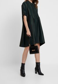 Abito oversize verde scuro con collo alto e maniche corte, caratterizzato da un corsetto aderente e un orlo svasato. Abbinato a stivaletti neri alla caviglia e una borsa con texture coccodrillo.