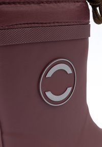 Bordó vízálló csizma texturált felsőrésszel, amelyen kör alakú fém logó található. A tetején húzózsinóros részlet.