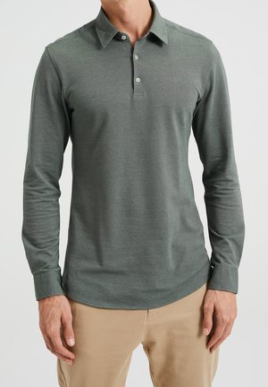 Mężczyzna ubrany w oliwkowo-zieloną koszulkę polo z długim rękawem i beżowe spodnie, stojący na tle jasnego, gładkiego tła.