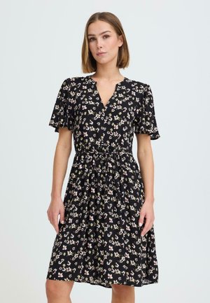 Woman wearing a black floral knee-length dress with short flutter sleeves and a gathered waist, standing against a plain light background.