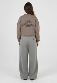 Brun oversized hettegenser med grafikk "Forbudt 10119 BERLIN" på ryggen, kombinert med grå vide bukser med teksturmønster.