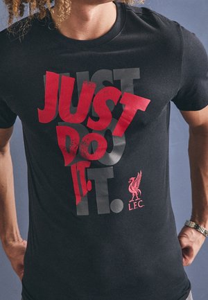 Mężczyzna noszący czarną koszulkę z czerwonym i ciemnoszarym napisem „JUST DO IT.” oraz małym czerwonym logo Liverpool FC w dolnej prawej części.