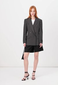 Grå stripete dobbeltrad blazer med store lapeller, paret med svarte shorts. Modellen har på seg svarte hæler med stropper og holder en liten veske.