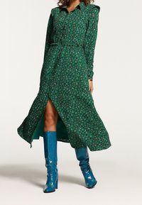 Grön blommig klänning med långa ärmar, knäppning framtill och en hög slits; kombinerad med glansiga blå knähöga stövlar.