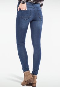 Jeans ajustados de color azul oscuro con una textura suave, que presentan dos bolsillos traseros, combinados con botas de tobillo de color marrón claro.