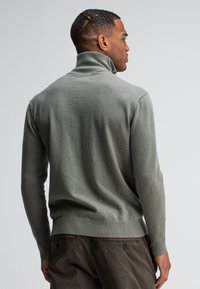 Groene gebreide trui met een hoge kraag, nauwsluitend ontwerp en lange mouwen. De textuur lijkt zacht, met een geribbelde onderkant en manchetten.