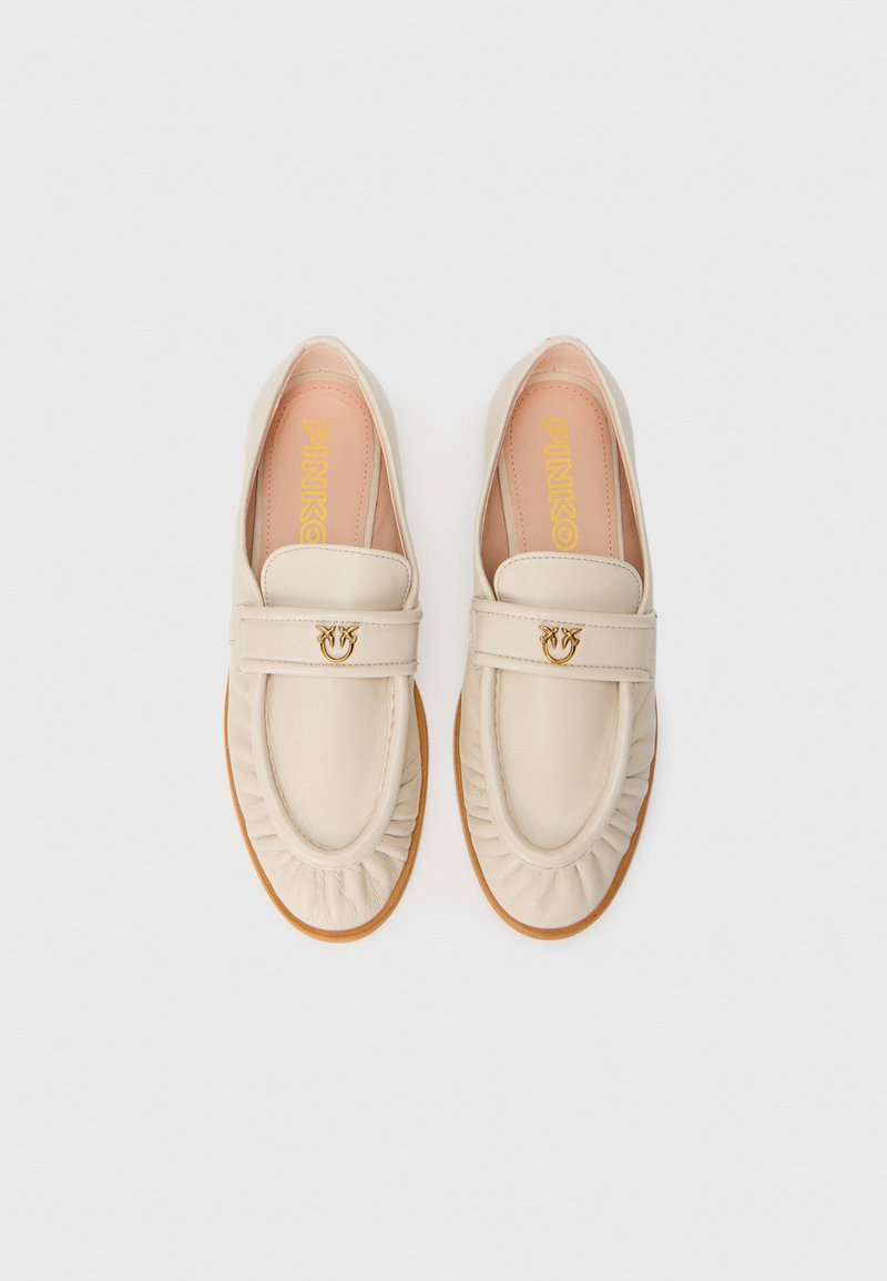 Mocasines de cuero beige con bordes ondulados, un detalle decorativo de hardware dorado en una correa y una suela de color claro. Textura suave y diseño clásico.