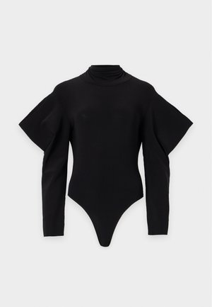 Femme portant un haut noir à manches longues avec des épaules bouffantes et un pantalon beige à jambes larges, debout devant un fond neutre.