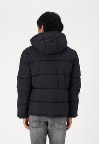 Svart polstret jakke med hette, med quiltede seksjoner og elastiske mansjetter. Plassert med ryggen vendt mot betrakteren.