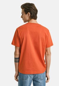 Kortærmet orange bomulds T-shirt med rund hals og en afslappet pasform, vist fra ryggen, parret med blå denim jeans.