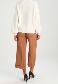 Weißer gerippter Strickpullover mit rundem Halsausschnitt; kombiniert mit hochgeschnittenen, weit geschnittenen braunen Hosen; schwarze High Heels.