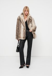 Abrigo de piel sintética en marrón claro y crema, combinado con una blusa negra y pantalones negros ajustados. La modelo lleva un pequeño bolso negro.