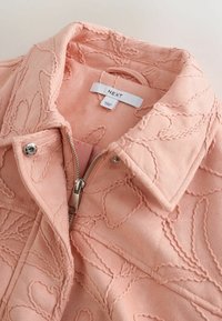 Veste rose pâle texturée avec motif floral brodé, fermeture éclair argentée et bouton-pression, col replié et étiquette de la marque "NEXT" visibles.