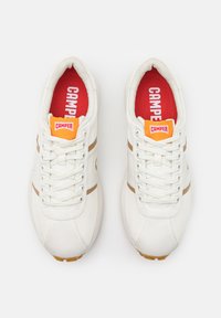 Λευκά αθλητικά παπούτσια με ανοιχτό καφέ ρίγες, υφή υλικού, στρογγυλή μύτη και κόκκινη σόλα που περιέχει τη λέξη "CAMPER" σε λευκό χρώμα.