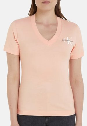 Femme portant un t-shirt Calvin Klein Jeans pêche clair à col en V avec manches courtes et pantalon foncé, cadrée au-dessus de la bouche.