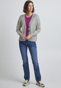 Cardigan en tricot gris avec un col rond et de grands boutons marron, superposé sur une chemise violette. Associé à un jean en denim bleu et des baskets.