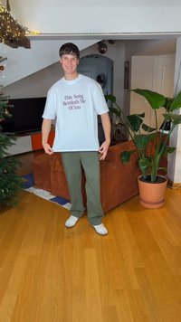 Πανύψηλο λευκό μπλουζάκι με μπορντό κείμενο «This Song Reminds Me Of You», συνδυασμένο με παντελόνι ελιάς και λευκά αθλητικά, σε εσωτερικό χώρου κατοικίας.