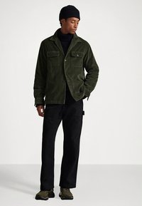 Ciemnozielona kurtka sztruksowa z dwoma przednimi kieszeniami, zestawiona z czarnymi spodniami, czarnym golfem i zielonymi sneakersami. Model pozuje na jednolitym tle.