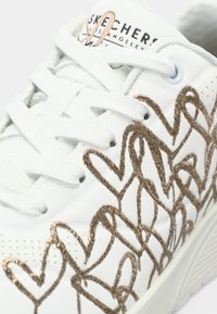 White sneaker with intricate gold heart-patterned stitching, round toe, and flat white laces, featuring a logo on the tongue.