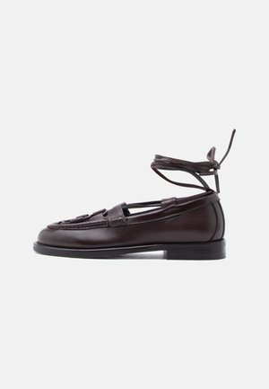 Tummanruskea nahkainen loafer, jossa nilkan ympärille kietoutuvat nauhat, matala korko ja punottu hihnallinen yksityiskohta jalan päällä.