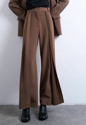 Brune bukser med vide ben og forreste plisseringer kombineret med sorte sko, båret med matchende brun blazer og sort top, stående på gråt gulv.