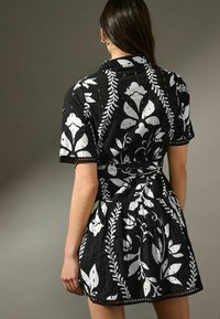 Robe noire avec un motif floral blanc et des accents de feuilles. Manches courtes, taille ceinturée, tissu texturé avec des détails en œillets et jupe évasée.