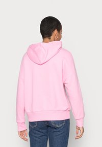 Sudadera con capucha rosa de corte holgado, mangas largas y puños acanalados. Tejido suave con diseño minimalista. Combinada con vaqueros azules.