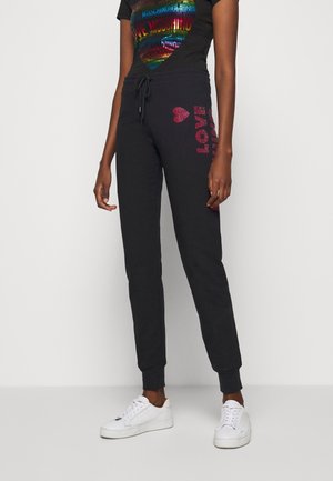 Femme portant un pantalon de survêtement noir avec texte rouge "LOVE" et motif cœur sur la cuisse, associé à des baskets blanches et un t-shirt graphique noir.