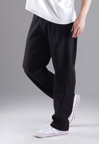 Schwarze Jeans mit einer lockeren Passform, seitlichen Taschen und einer glatten Textur, kombiniert mit weißen Low-Top-Sneakern mit roten Akzenten.