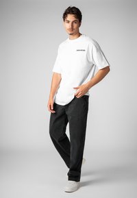 Weißes Baumwoll-T-Shirt mit schwarzem Aufdruck "WORLDWIDE", kombiniert mit schwarzer Jeans aus Denim. Das Modell steht mit den Händen in den Taschen und weißen Sneakers.