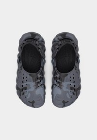 Ζευγάρι από σκούρα γκρι και μαύρα παπούτσια κλογκ με μοτίβο καμουφλάζ, με οπές αερισμού και ρυθμιζόμενα λουριά στη φτέρνα, φωτογραφημένα από πάνω σε λευκό φόντο.