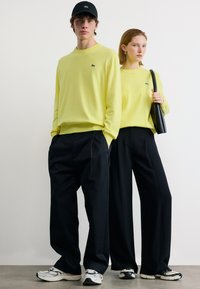 Νεαρός άνδρας και γυναίκα φορούν ταιριαστά κίτρινα φούτερ, μαύρο παντελόνι φαρδύ, και λευκά αθλητικά παπούτσια, ποζάροντας μπροστά σε απλό λευκό φόντο.