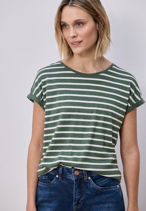 Kvinde med kort blondt hår iført en grøn og hvid stribet t-shirt med opkavlede ærmer, stoppet ned i blå jeans, der kigger mod højre.