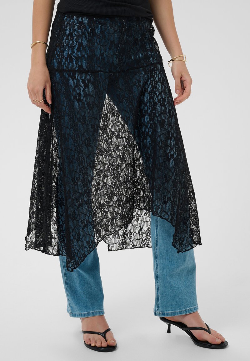 Femme portant une sur-jupe asymétrique en dentelle noire associée à un jean bleu et des sandales noires à talons, montrée de la taille aux pieds.